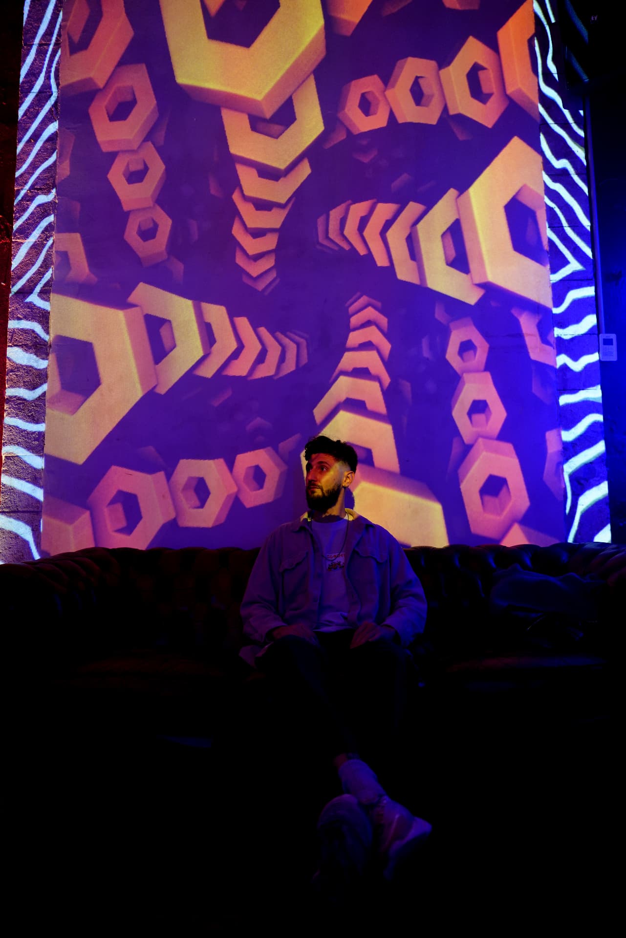 A person seated on a leather sofa looking upward, behind them a projection of yellow prisms in perspective against a blue backdrop