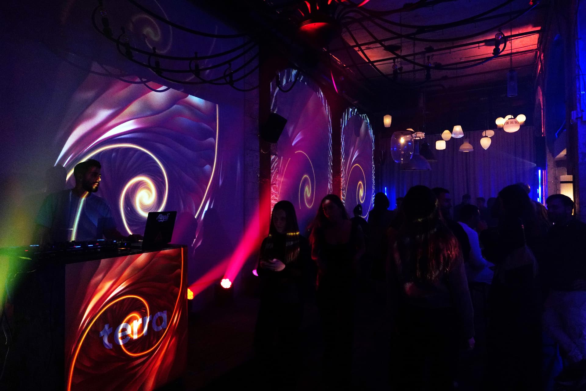 A DJ at a lit booth marked terra, with spiral projections across a brick wall and arched alcoves, guests on the floor