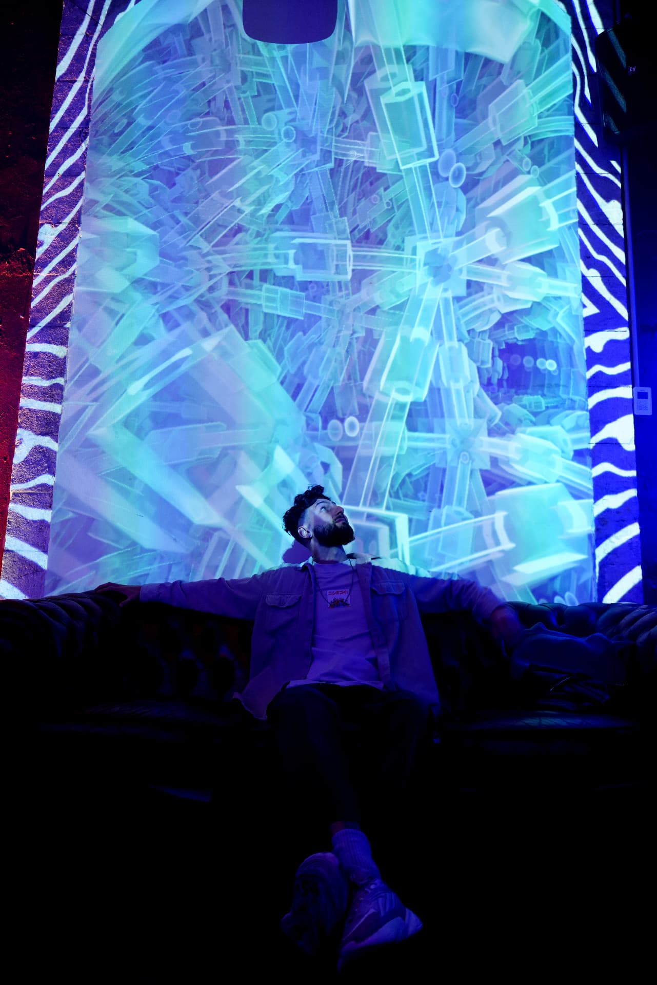 A person seated on a leather sofa looking upward, behind them a large arch projected with black-and-white stripes and a lattice pattern