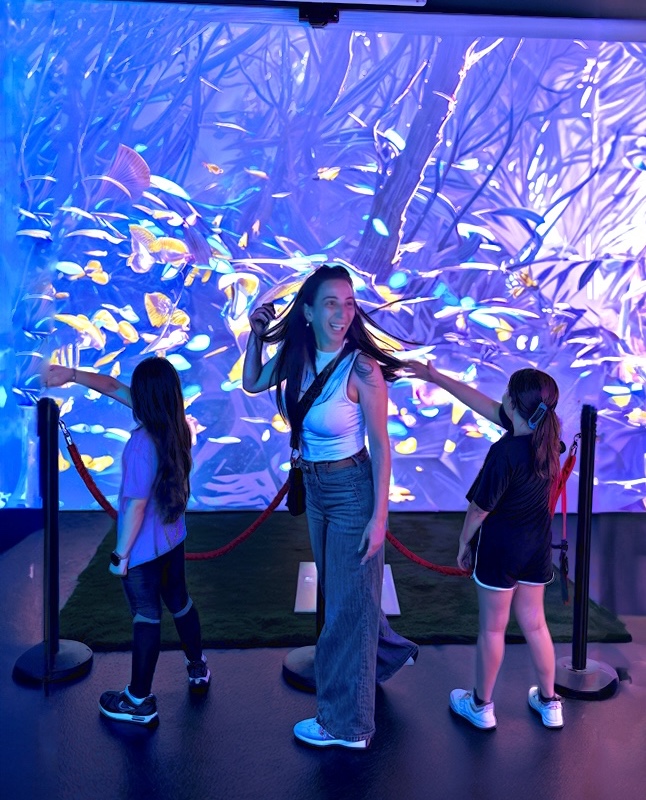 A woman and two girls in front of a colorful jungle projection with butterflies in the Bugs World installation
