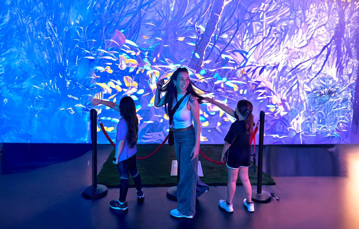 A woman and two girls in front of a colorful jungle projection with butterflies in the Bugs World installation