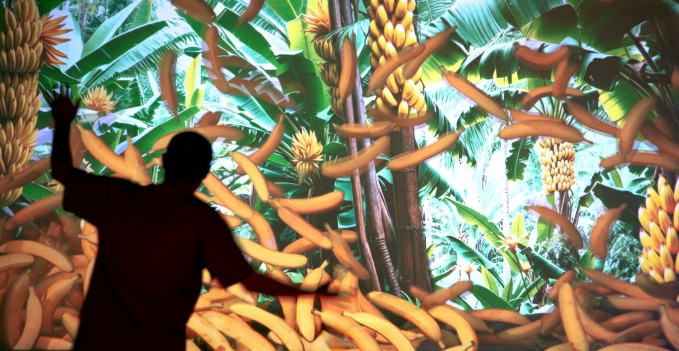 A visitor raises his hands in front of a tropical forest and banana projection in the Interactive Jungle installation